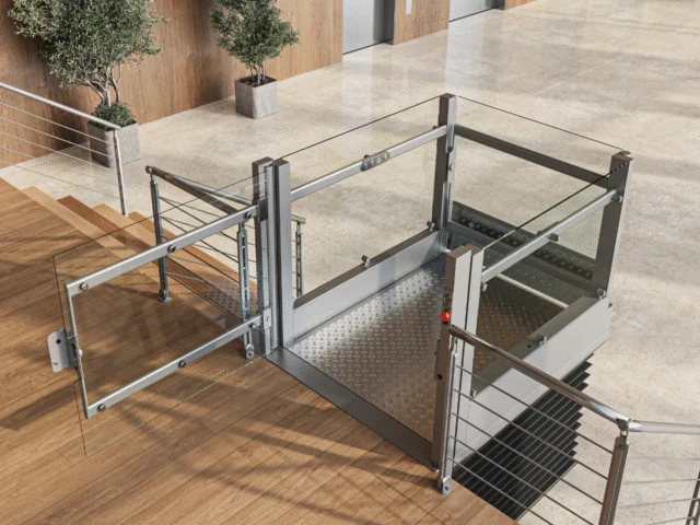 A modern wheelchair lift with glass panels is installed next to a staircase in an indoor public building with elevators and potted plants.