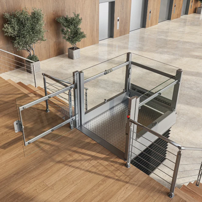 A modern wheelchair lift with glass panels is installed next to a staircase in an indoor public building with elevators and potted plants.