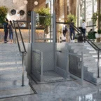 A wheelchair lift is installed between two marble staircases in a hotel lobby, with people conversing near the reception desk in the background.