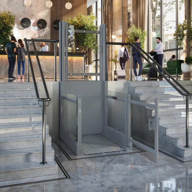 A wheelchair lift is installed between two marble staircases in a hotel lobby, with people conversing near the reception desk in the background.