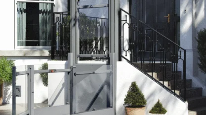 A residential wheelchair lift is installed outside a house, next to a staircase leading to the front door, with potted plants nearby.