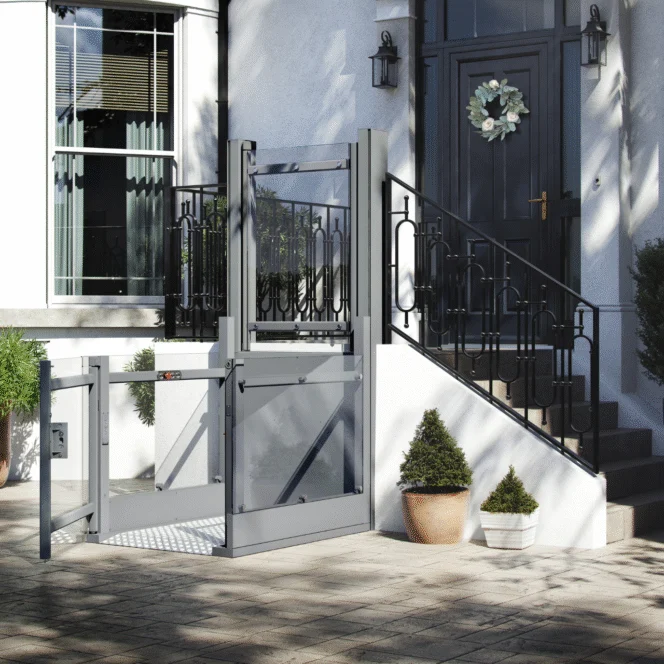 A residential wheelchair lift is installed outside a house, next to a staircase leading to the front door, with potted plants nearby.