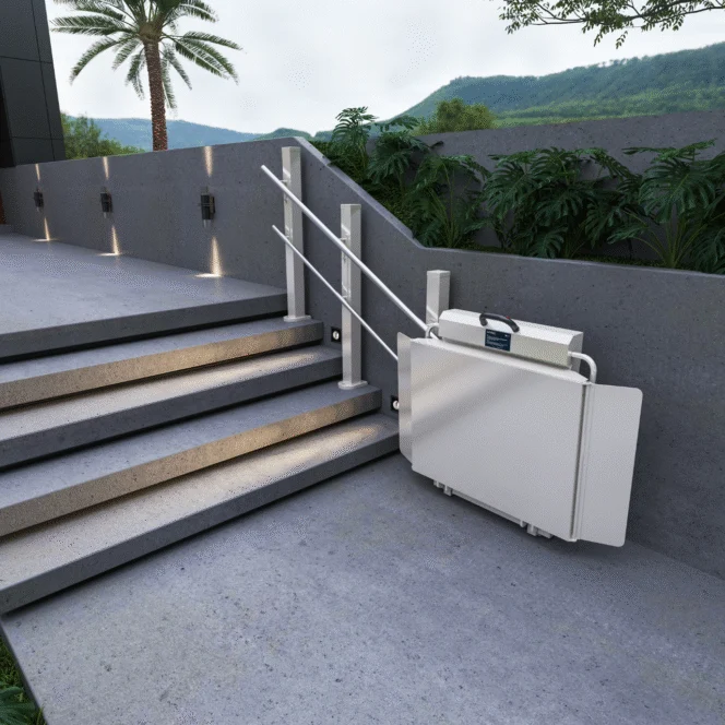 A folded platform lift is installed beside outdoor stairs with a handrail, next to a wall and greenery, under a cloudy sky.