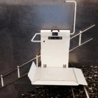 A stair lift platform for wheelchair users installed on a staircase with metal railings, against a gray wall.