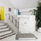 A wheelchair platform lift is installed next to a staircase for accessibility in a white-tiled indoor space with a yellow door and potted plant visible.