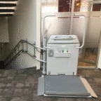 A stairlift platform for wheelchair users is installed next to a staircase in an indoor setting with carpeted floors and glass partition walls.