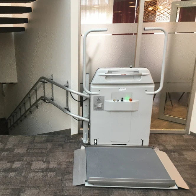 A stairlift platform for wheelchair users is installed next to a staircase in an indoor setting with carpeted floors and glass partition walls.