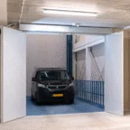 A black Peugeot van is parked inside a well-lit, industrial-style indoor garage with open double doors and blue railings on one side.