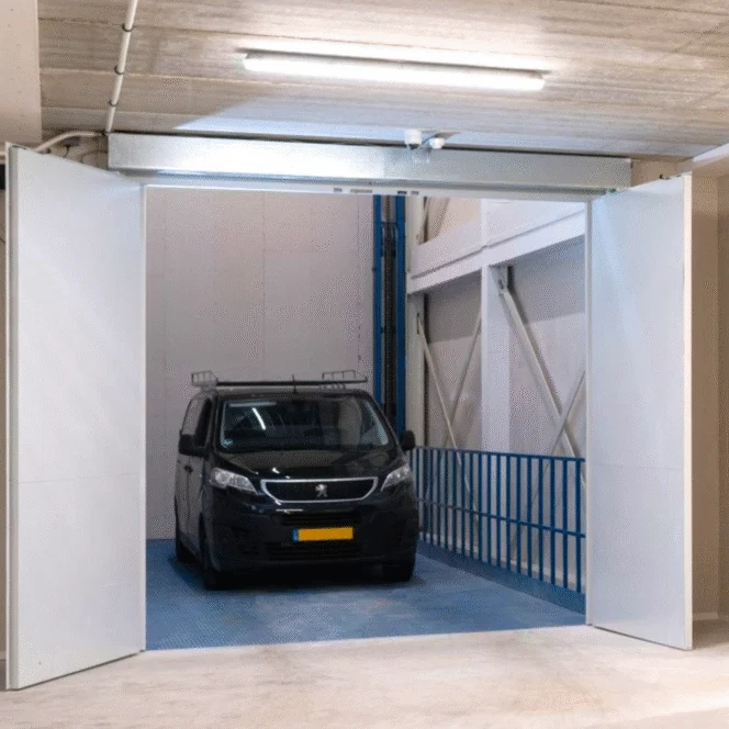 A black Peugeot van is parked inside a well-lit, industrial-style indoor garage with open double doors and blue railings on one side.