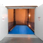 A rectangular indoor swimming pool with a blue cover, surrounded by metal railings and illuminated by warm lighting in an industrial-style room.