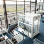 A modern glass elevator is located beside a staircase in a large, bright indoor space overlooking a racetrack through tall windows.