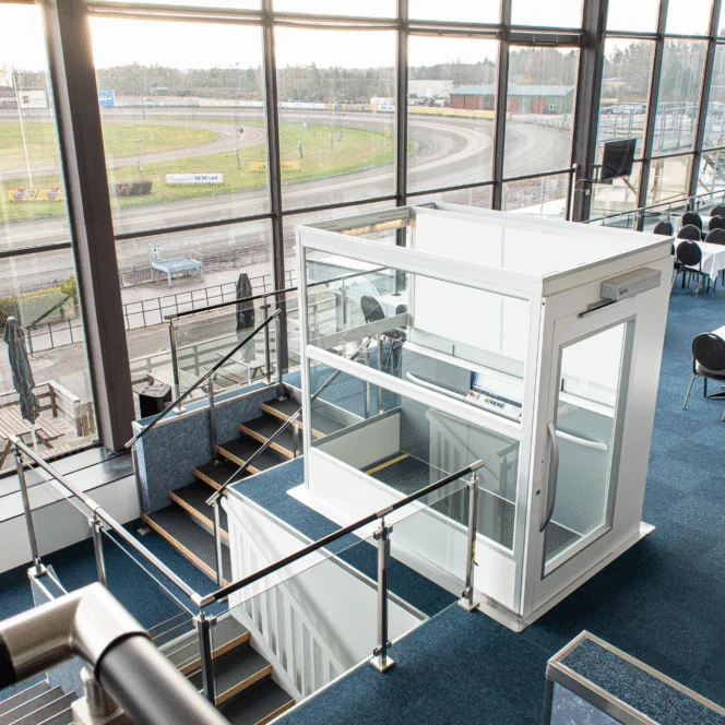 A modern glass elevator is located beside a staircase in a large, bright indoor space overlooking a racetrack through tall windows.
