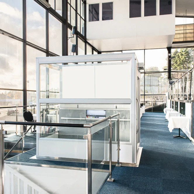 A modern, glass-enclosed office booth with a desk and chair inside, located in a spacious, sunlit building with large windows and blue carpeted floor.