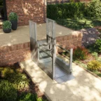 A metal wheelchair lift is installed next to a brick patio, providing accessible entry from a garden path to the raised outdoor area.