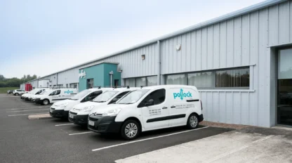 Several white company vans are parked in a row outside a large industrial warehouse building on a clear day.