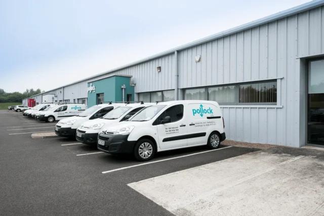 Several white company vans are parked in a row outside a large industrial warehouse building on a clear day.