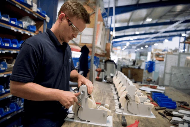 A man wearing safety glasses assembles mechanical parts at a workbench in a factory, with rows of components and tools organized on shelves and the table.