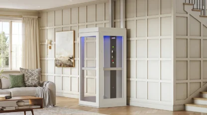 A compact home elevator with glass panels and control buttons is installed in a modern living room with paneled walls and wood flooring.