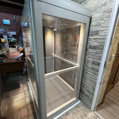 A small, modern glass elevator with white railings inside, positioned next to a stone-textured wall and tile flooring in an indoor setting.