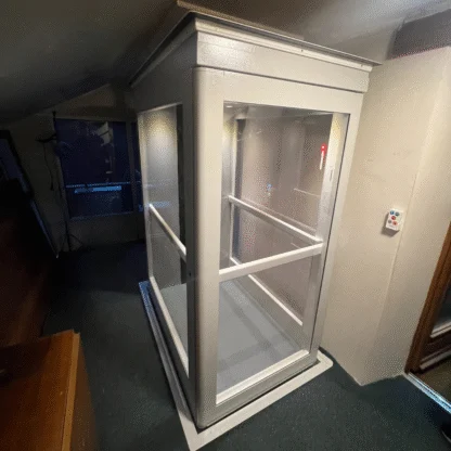 A small glass residential elevator with white framing installed indoors, showing a closed door and control panel on the wall outside.