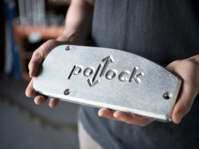 A person holds a rectangular metal plate embossed with the word "Pollock" and two arrows pointing up and down.