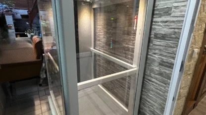 A small, glass-enclosed indoor elevator with metal frame, positioned next to a stone-textured wall in a modern interior space.