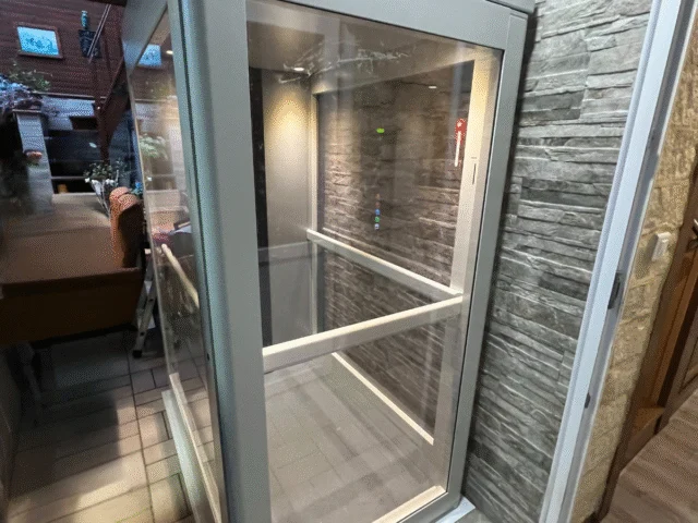 A small, glass-enclosed indoor elevator with metal frame, positioned next to a stone-textured wall in a modern interior space.