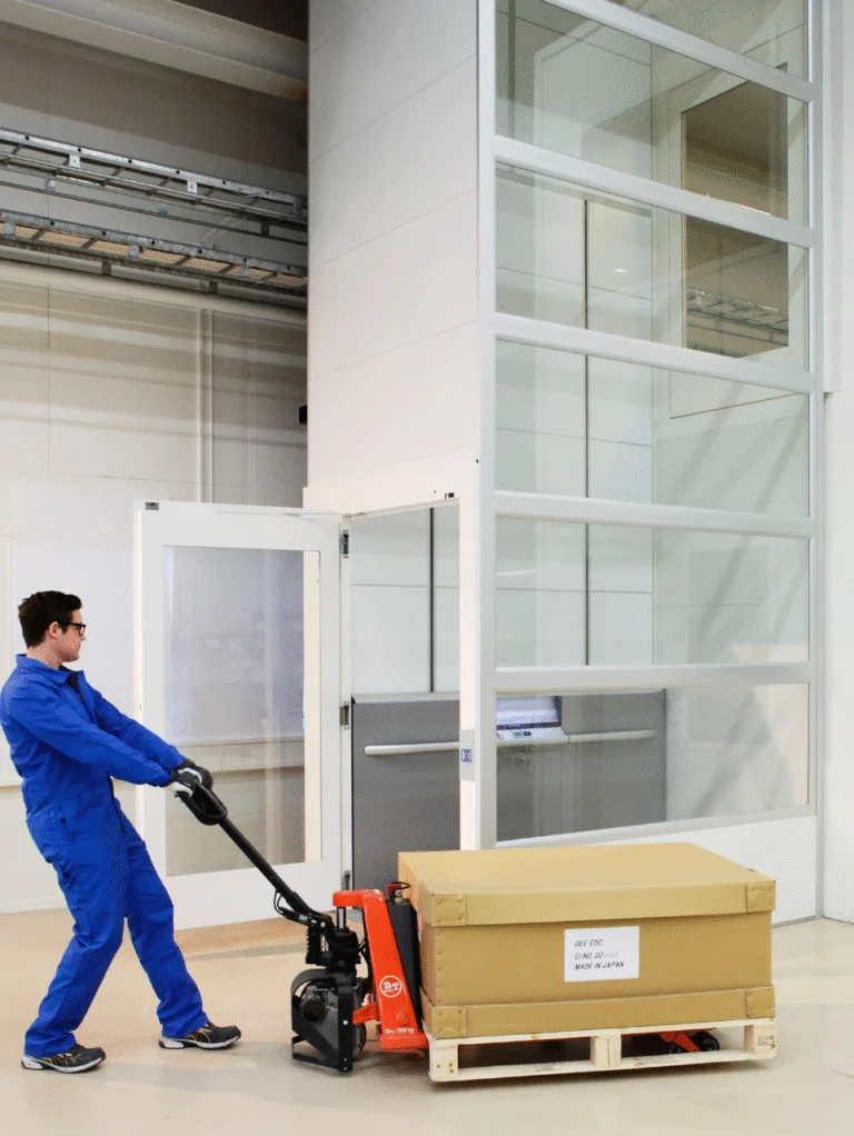 A person in blue coveralls uses a pallet jack to move a large cardboard box on a wooden pallet through an open door in an industrial setting.