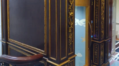 A small modern glass elevator with ornate dark wood and gold decorative paneling on its exterior, located inside a building near a staircase with a decorative railing.