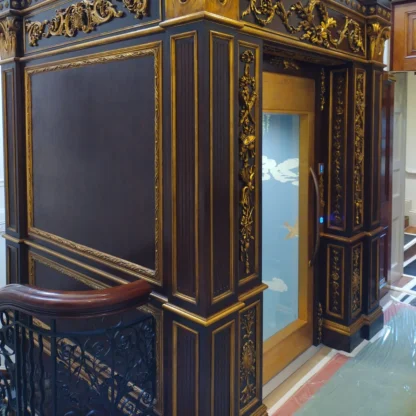 A small modern glass elevator with ornate dark wood and gold decorative paneling on its exterior, located inside a building near a staircase with a decorative railing.