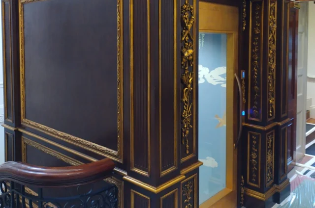 A small modern glass elevator with ornate dark wood and gold decorative paneling on its exterior, located inside a building near a staircase with a decorative railing.