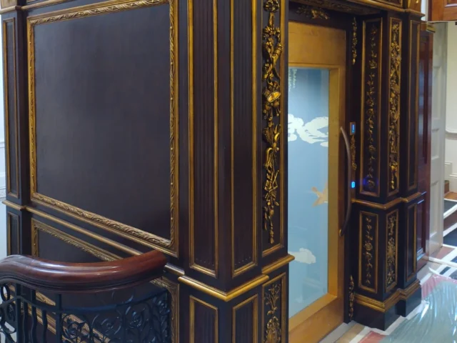 A small modern glass elevator with ornate dark wood and gold decorative paneling on its exterior, located inside a building near a staircase with a decorative railing.