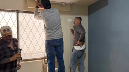 Three men work together in a room; one stands on a ladder adjusting a ceiling panel, while the others assist on the ground near tools and a window.