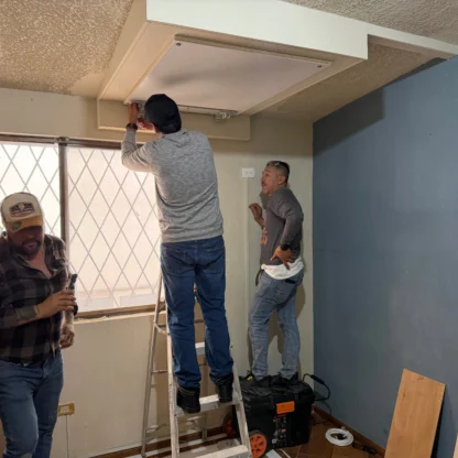 Three men work together in a room; one stands on a ladder adjusting a ceiling panel, while the others assist on the ground near tools and a window.