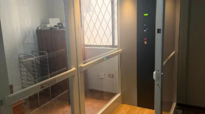 A small residential elevator with glass walls, wood-patterned floor, and control panel, located indoors near a metal rack and furniture.