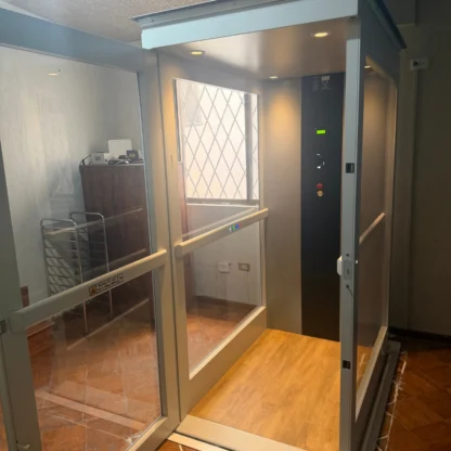 A small residential elevator with glass walls, wood-patterned floor, and control panel, located indoors near a metal rack and furniture.