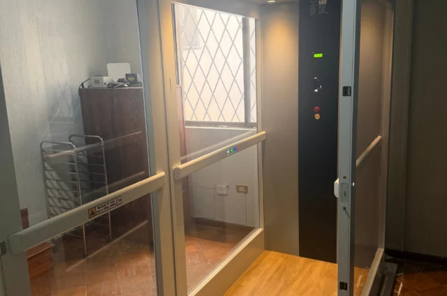 A small residential elevator with glass walls, wood-patterned floor, and control panel, located indoors near a metal rack and furniture.