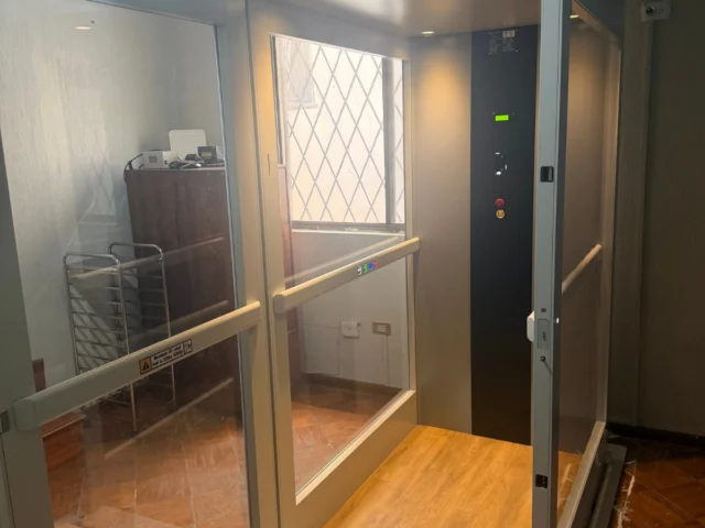A small residential elevator with glass walls, wood-patterned floor, and control panel, located indoors near a metal rack and furniture.