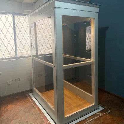 A small, glass-walled elevator with a metal frame is installed indoors on a wooden floor, next to a window with a diamond-patterned grille.