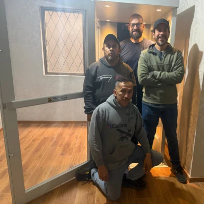 Four men posing inside and in front of an open elevator, with wooden flooring and patterned wallpaper visible in the background.