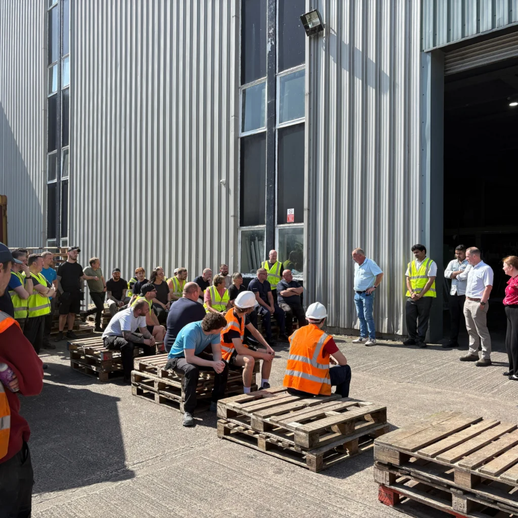 A group of people, some in safety vests and helmets, gathered outside a large industrial building, sitting and standing on wooden pallets during daylight.
