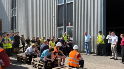 A group of people, some in safety vests and helmets, gathered outside a large industrial building, sitting and standing on wooden pallets during daylight.