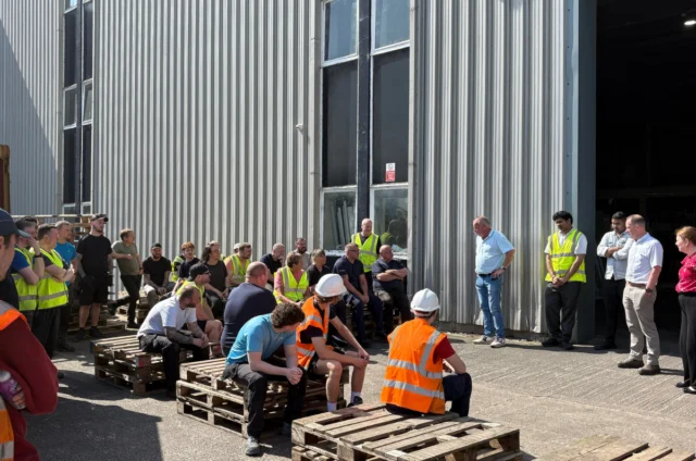 A group of people, some in safety vests and helmets, gathered outside a large industrial building, sitting and standing on wooden pallets during daylight.