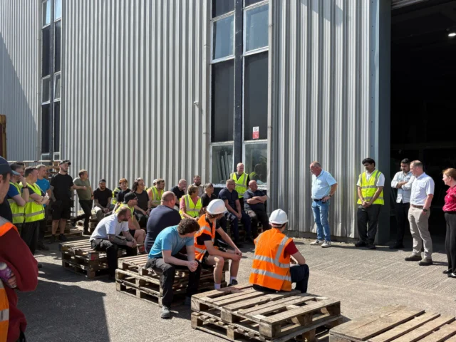 A group of people, some in safety vests and helmets, gathered outside a large industrial building, sitting and standing on wooden pallets during daylight.