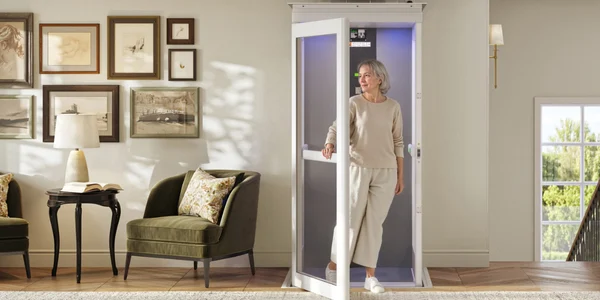 Older woman exits a small residential elevator in a modern, well-lit living room with framed wall art, armchairs, and a side table.