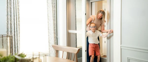 A woman helps a young boy with glasses exit a home elevator into a bright dining room.
